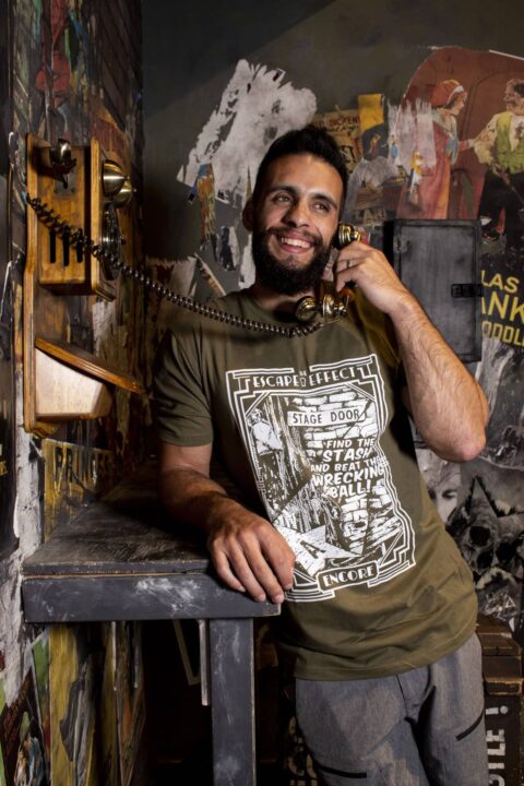 Encore Shirt Model, Green Variant 6 A man is talking on a telephone, wearing a green "Encore" shirt featuring an illustration of the backstage area of an abandoned theater.
