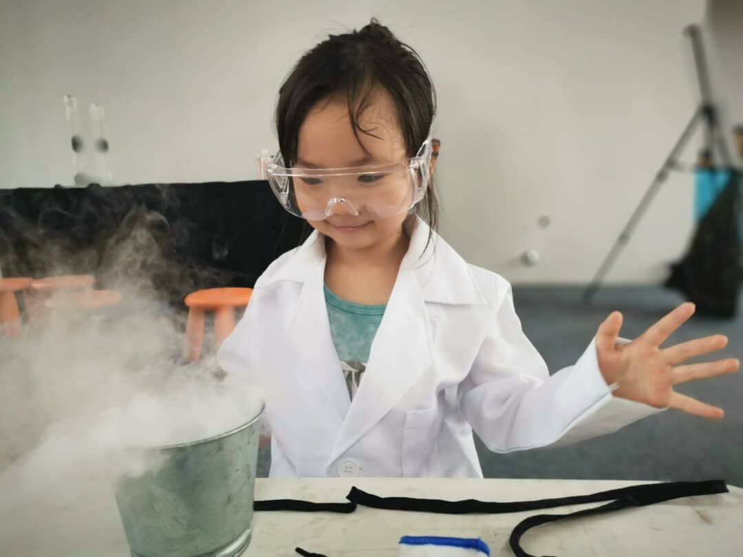 A kid in a lab coat and safety glasses performing an experiment in a science center