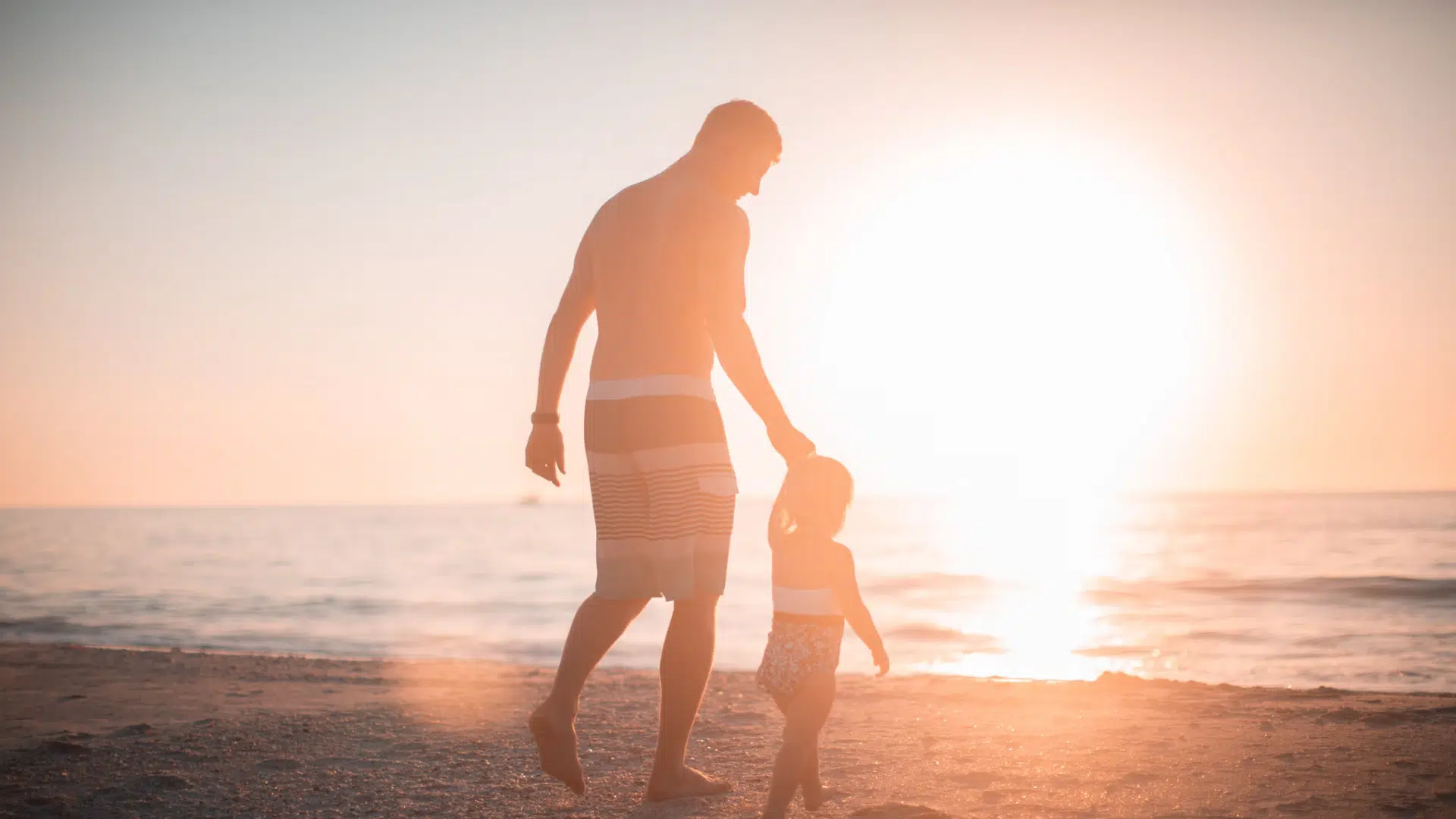 A father and his child walking on a beach at sunset.