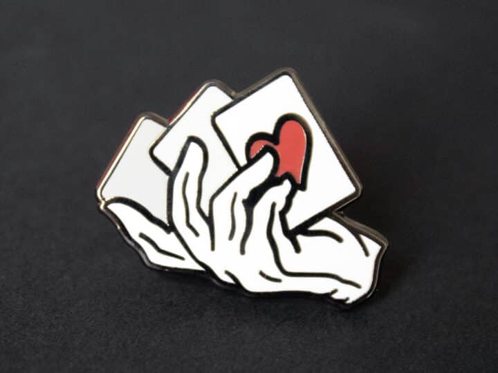 A close up of a magician's hand and cards lapel pin.