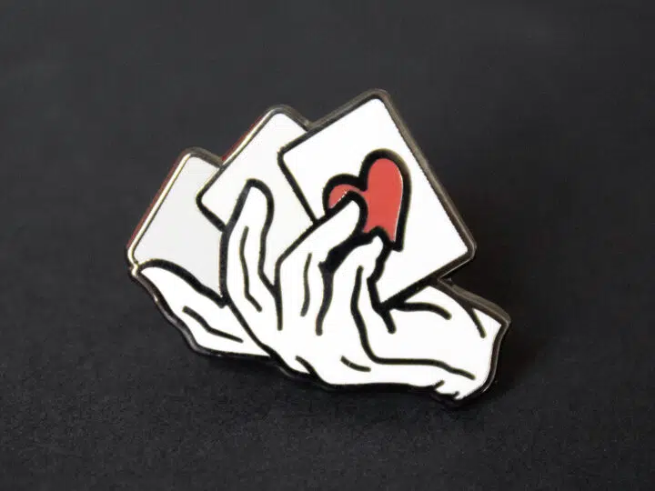 A close up of a magician's hand and cards lapel pin.