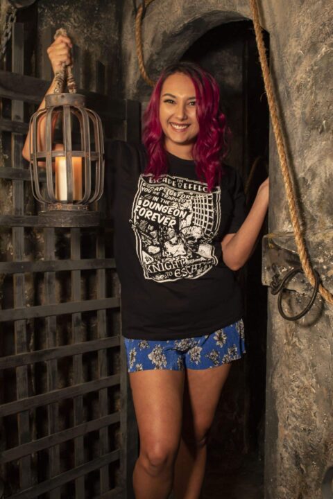 Female model poses in the dungeon of "A Knight To Escape" with a black graphic tee shirt.