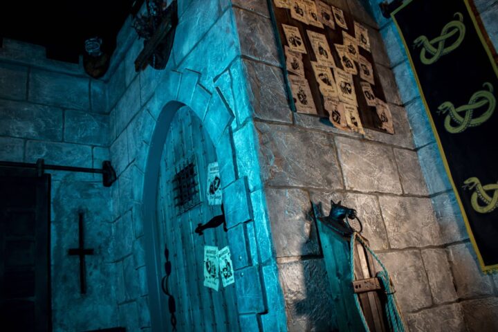 An outside view of the stone walls of the castle decorated with a banner, a notice board covered with wanted posters and a hitching post.