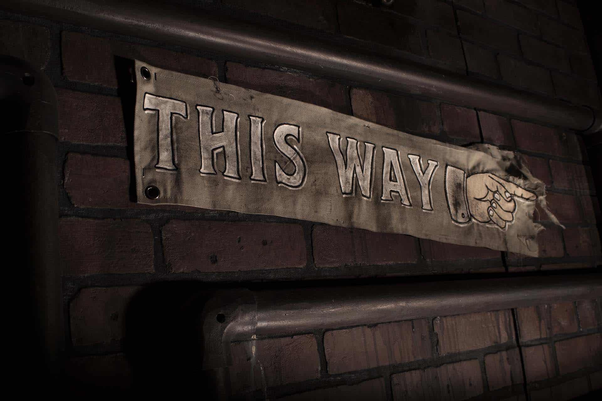 A photograph of a distressed banner with the words This Way written across it and a pointing hand.