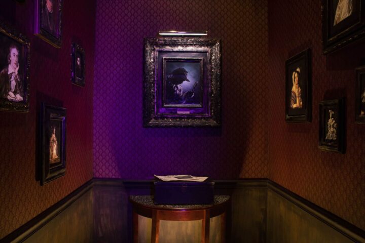 A plain wooden box sits on a table, littered with newspapers, underneath a portrait of Sherlock Holmes.